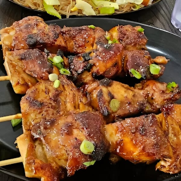 Grilled Filipino chicken skewers, known as chicken inasal, garnished with green onions on a black plate.