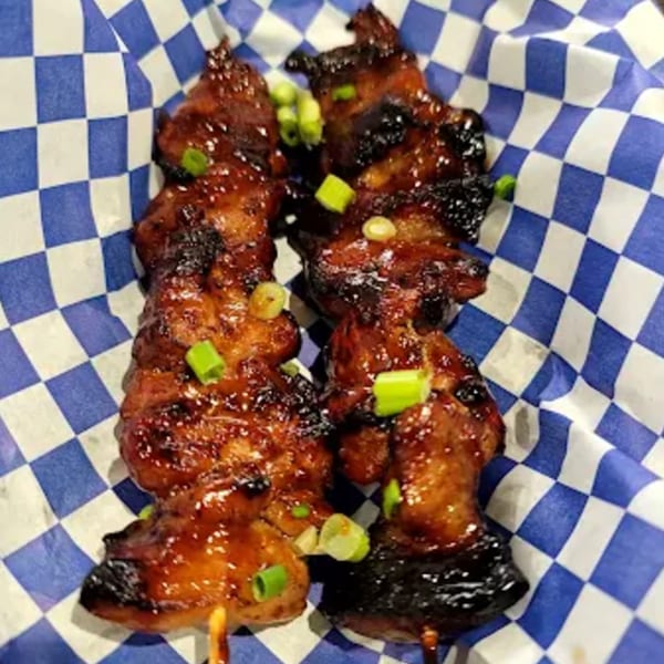 Two skewers of glistening Filipino BBQ pork, garnished with green onions, in a blue and white checkered basket.