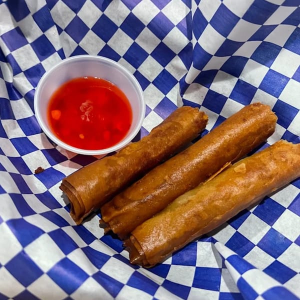 Three golden-brown Filipino lumpia spring rolls with a side of red dipping sauce on blue and white checkered paper.