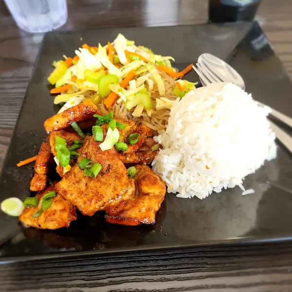 Filipino adobo chicken with rice, pancit, and mixed vegetables on a black plate.