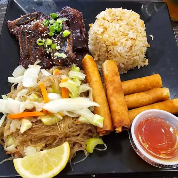 Filipino meal with BBQ ribs, fried rice, pancit, lumpia, and a lemon wedge on a black plate.